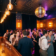Dunkel beleuchtete Bar mit Edison-Glühbirnen, Discokugel streut Licht in Amber- und Violetttönen über Cocktailgläser und Tanzfläche.