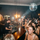 Belebte Bar mit Disco-Spiegelkugel, Edison-Glühbirnen und tanzender Menge in warmem Amber- und Silberlicht.