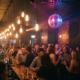 Belebte Bar mit Diskokugel, Edison-Glühbirnen und feiernden Gästen in magentafarbenem Licht vor Backsteinwand.