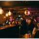 Belebte Bar mit Dancefloor, Edison-Glühbirnen und Spiegelkugel tauchen Silhouetten junger Menschen in warmes Gold und Pink.
