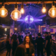 Belebte Bar-Tanzfläche mit Discokugel, Edison-Glühbirnen in Bernsteinton und violetten Neonakzenten über tanzenden Gästen.