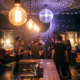 Belebte Bar mit Tanzfläche, Edison-Glühbirnen und Diskokugel tauchen Silhouetten junger Gäste in Burgund- und Ambertöne.