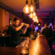 Junger Bartender schüttelt Cocktail-Shaker an elegantem Tresen in einem Münchner Nachtclub mit warmem Edison-Licht.