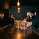 Bartender reicht zwei beschlagene Cocktails über einen dunklen Bartresen, warme Edison-Glühbirnen und verschwommene Gäste im Hintergrund.