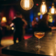Cocktailglas mit bernsteinfarbenem Licht auf dunkler Marmorbar, Edison-Glühbirnen und verschwommene Tanzfläche im Hintergrund.