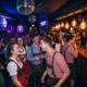 Junge Feiernde in Dirndl und Lederhosen tanzen in einem Nachtclub mit Diskokugeln und Edison-Glühbirnen.
