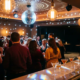 Discokugel wirft Lichtreflexe auf eine belebte Tanzfläche mit warmem Ambiente, Edison-Glühbirnen und Cocktailgläsern in Burgund- und Goldtönen.