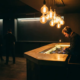 DJ an beleuchteter Bar-Konsole, Edison-Glühbirnen tauchen leere Tanzfläche in warmes Bernsteinlicht, Silhouetten im Hintergrund.