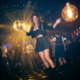 Junge Frau im modernen Dirndl tanzt ausgelassen auf dunkler Clubbühne mit Discokugel-Reflexionen und warmem Ambiente-Licht.