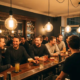 Gäste lachen bei Craft Cocktails an einer warmbeleuchteten Theke in einer Münchner Bar mit Edison-Glühbirnen und dunklem Holz.
