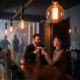 Zwei lachende Gäste mit Cocktails an einer stilvollen Bar in München, Edison-Glühbirnen, warmes Ambiente, Tanzfläche im Hintergrund.