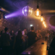 Münchner Bar mit Discokugel, die farbiges Licht auf Backsteinwände wirft, Edison-Glühbirnen und tanzende Silhouetten im Hintergrund.