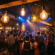 Münchner Bar mit Tanzfläche, Discokugel und Edison-Glühbirnen, junge Feiernde in Oktoberfest-Nightclub-Outfits, Backsteinwände in Amber- und Blautönen.