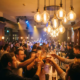 Belebte Münchner Bar mit Edison-Glühbirnen, erhobenen Cocktailgläsern und Tanzfläche in warmem Bernsteinlicht.