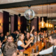 Diskokugel wirft Lichtreflexe in einer belebten Münchner Bar, Gäste stoßen lachend mit Cocktails an unter warmem Glühlampenlicht.