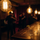 Münchner Bar mit Tanzfläche in warmem Bernsteinlicht, Edison-Glühbirnen, leeres Cocktailglas auf dem Tresen, silhouettierte Figuren im Hintergrund.
