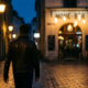 Person in stilvoller Jacke auf nassen Münchner Kopfsteinpflaster-Straßen bei Nacht, warme Bernstein-Straßenlaternen, Bar mit Edison-Glühbirnen im Hintergrund.