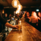Bartender reicht einen Cocktail über eine glänzende Theke in einer Münchner Bar mit Edison-Glühbirnen und belebter Tanzfläche im Hintergrund.