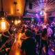 Junge Feiernde tanzen unter einer Diskokugel in einer stimmungsvollen Bar mit Edison-Glühbirnen und violettem Akzentlicht.