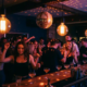 Lebhafte Tanzfläche in einer Bar mit Edison-Glühbirnen, Discokugel und Cocktailgläsern in warmen Amber- und Magenta-Tönen.