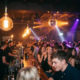 Belebte Bar mit Tanzfläche, Edison-Glühbirnen und Diskokugel in Bernstein- und Violetttönen.