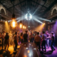 Tanzende Menge in einer Bar mit Diskokugel, Edison-Glühbirnen und violett-bernsteinfarbenem Licht in industriellem Ambiente.