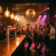 Junge Feiernde tanzen unter einer Diskokugel in einer stimmungsvollen Bar mit Edison-Glühbirnen und Lichtreflexen in Amber und Magenta.