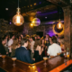 Belebte Bar mit tanzender Menge, Edison-Glühbirnen und Diskokugel, die goldenes Licht an dunkle Wände wirft.