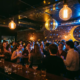 Belebte Bar mit Discokugel, die goldenes und blaues Licht auf Backsteinwände und Cocktailgläser wirft, Edison-Lampen erhellen die Tanzfläche.