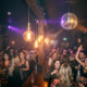 Belebte Bar-Tanzfläche mit Discokugel, Edison-Glühbirnen und feiernden Gästen in warmem Bernstein- und Violettlicht.