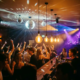 Tanzende Menge in belebter Bar mit Discokugeln, Edison-Glühbirnen und farbigen DJ-Lichtern in Lila und Amber.