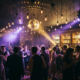 Stimmungsvolle Bar mit Tanzfläche, beleuchtet von Edison-Glühbirnen, Discokugel-Reflexionen und violetten Lichtstrahlen in goldener Atmosphäre.