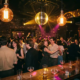 Tanzende Menschenmenge unter Discokugel und Edison-Glühbirnen in einer stimmungsvollen Bar mit goldenem und violettem Licht.