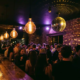 Belebte Tanzfläche einer Bar mit Edison-Glühbirnen, Discokugel und warmem Licht in Bernstein- und Lilatönen.