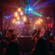 Vollbesetzte Bar mit Tanzfläche, Edison-Glühbirnen und Discokugel, Silhouetten tanzender Menschen in warmem Bernstein- und Neonlicht.