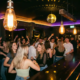 Belebte Bar mit Dancefloor, Edison-Glühbirnen und Diskokugel tauchen junge Menge in goldenes und violettes Licht.