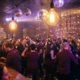 Tanzende Menge in belebter Bar mit Edison-Glühbirnen, Discokugeln und stimmungsvollem Licht in Burgund, Gold und Violett.