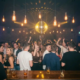 Feiernde Menschen auf einer Tanzfläche unter einer Discokugel mit goldenem Licht, Edison-Glühbirnen und Cocktails an der Bar.