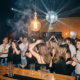 Belebte Bar mit Discokugel, Vintage-Edison-Glühbirnen und tanzenden Gästen in warmem Bernstein- und Marinblaulicht.