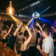 Tanzende Menge unter Discokugel und Edison-Lampen in einer stimmungsvollen Bar mit Backsteinwänden und Cocktails auf dem Tresen.