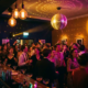 Feiernde Menge in einer stimmungsvollen Bar mit Edison-Glühbirnen, Discokugel und bunten Cocktailgläsern in Navy und Gold.