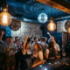 Tanzende Menge in Oktoberfest-Outfits unter Edison-Glühbirnen und Diskokugel in einer stimmungsvollen Bar.