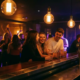 Studierende lachen an einer modernen Bar, Edison-Glühbirnen tauchen den Raum in warmes Amber- und Violettlicht, Tanzfläche im Hintergrund.