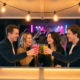 Junge Studierende stoßen lachend mit bunten Cocktails an einer beleuchteten Bar an, warmes Ambiente mit Edison-Glühbirnen und Tanzfläche im Hintergrund.