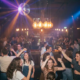 Studierende tanzen ausgelassen unter einer Diskokugel in einer stimmungsvollen Bar mit Edison-Lampen und farbigen Bühnenlichtern.