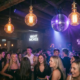 Belebte Bar-Tanzfläche mit Edison-Glühbirnen und Discokugel, die warmes Bernstein- und Violettlicht über tanzende Gäste streut.