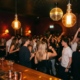 Tanzende Menge unter Disco-Kugel und Edison-Glühbirnen in einer stimmungsvollen Bar mit Cocktails im Amber-Licht.