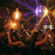 Tanzende Gäste in einer Bar mit Discokugel, Edison-Glühbirnen und warmem Licht aus Gold- und Lilatönen auf Backsteinwänden.