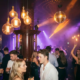 Disco-Kugel verstreut warmes Licht über tanzende Gäste in einer Bar mit Edison-Lampen, lila und bernsteinfarbenen Lichtstrahlen und Nebeleffekten.