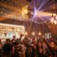 Glitzernde Diskokugel wirft Lichtreflexe über tanzende Gäste mit Getränken unter warmen Edison-Glühbirnen in lebhafter Nachtclub-Atmosphäre.